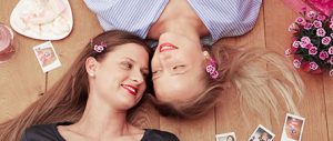 Zwei Freundinnen mit Pink Kisses Blüten im Haar liegen auf dem Boden und lächeln sich an