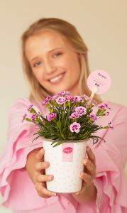 Eine junge Frau hält dir lächelnd eine Pink Kisses Mininelke im Übertopf mit lauter kleinen rosafarbenen Kussmündern daruaf entgegen. Auf der Rückseite des in der Pflanze steckenden Frienstick-Pflanzensteckers steht "For you". Für eine personalisierte Unterschrift ist eine gestrichelte Linie darunter.